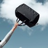 A black duffle bag is being held up against a blue sky with clouds. The bag features dual handles and a logo.