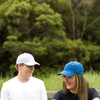 A blue and a white cap are worn by two people outdoors, surrounded by greenery. Each cap has a logo.