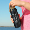 A drink bottle in matte black with a wooden top and a branding logo, held against a scenic background.