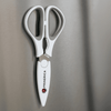 Kitchen scissors with a white handle and magnetic holder, mounted on a metallic surface. Features a logo.
