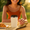 A white double wall coffee cup with a black lid sits on a table beside a croissant, with a smiling person in the background.