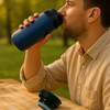 A drink bottle in navy blue held by a man outdoors, with a lid resting on a table nearby.