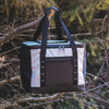 A cooler bag in grey and black with sturdy handles and a front pocket, hanging from a tree branch.