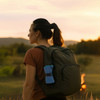 A woman with a ponytail wearing a brown top and carrying a green backpack, set against a sunset landscape.