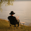 A person in a black hat sits in a black folding chair, reading a book beside a small table by the water's edge.