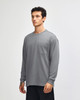 A man is wearing a grey long-sleeve t-shirt with a pocket, standing in a neutral backdrop. The shirt has a logo.
