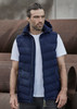 A man wearing a navy blue unisex hooded puffer vest over a white t-shirt, standing in an industrial setting.