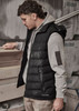 A black unisex hooded puffer vest worn over a grey sweatshirt, featuring a small logo on the vest.