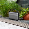 A premium wireless speaker in silver and black, positioned among greenery on a rocky surface.