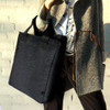 A black tote bag with sturdy handles, held by a person in a grey coat. The bag features a simple, modern design.