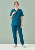 A model wearing a teal women's scrub top and matching pants, featuring a logo, against a light background.