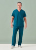A man wearing unisex teal scrub pants and a matching scrub top, standing against a light green background.