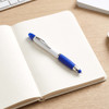 A blue and white ballpoint pen rests on an open notebook with lined pages. The background includes a keyboard and a coffee cup.