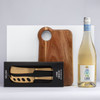 A wooden cheese board with a handle, alongside a gold 2-piece cheese knife set and a white wine bottle.