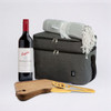A grey cooler bag with a red wine bottle, a grey blanket, and a wooden cheese board with a gold cheese knife.