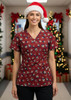 A women's Christmas scrub top in burgundy with festive patterns, worn by a smiling model in a Santa hat.