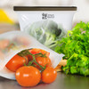 A silicone reusable storage pouch in transparent white containing broccoli, with fresh tomatoes and parsley beside it.