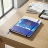 A full colour blue notebook with a logo, placed on a wooden table alongside papers and a tablet.