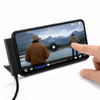 A smartphone on a stand showing a video with a person touching the screen. The phone is connected with a charging cable.