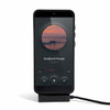 A black phone on a stand displaying a music player interface with a charging cable attached.