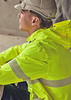 A man in a grey helmet wears a bright yellow rain jacket with reflective stripes, seated in a construction environment.