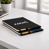 A black and navy A4 notebook with a logo, stacked on a desk next to a small pot plant.