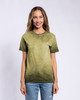 A woman stands smiling in an olive green Colortone® Oil Wash T-Shirt, paired with light blue jeans. The shirt has a branding logo.