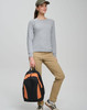 A woman wearing a grey top and khaki pants holds a black and orange Iconic Promo Backpack.