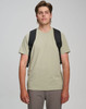 A man wearing a light khaki T-shirt and grey pants, carrying a black backpack with shoulder straps.