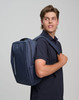 A navy blue backpack is worn over one shoulder by a young man in a navy polo shirt. The backpack features a logo.
