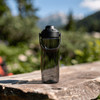 A drink bottle in a sleek, transparent design with a black lid and handle, set against a natural outdoor backdrop.