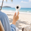A white rechargeable fan is held by a person, set against a beach background with sand and palm trees.