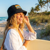 A woman wearing a black terry towelling bucket hat with a full-colour patch, smiling on a beach with palm trees.