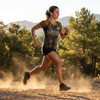 A woman runs on a dirt path, wearing a yellow and black active V-neck t-shirt and shorts, surrounded by trees and mountains.