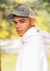 A man in a grey cap and white hoodie stands outdoors, showcasing a stylish look against a blurred natural background.