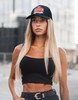 A woman with long blonde hair wearing a black tank top and cap with a logo stands outdoors.