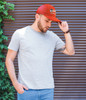 A man wearing a grey t-shirt and a red cap stands against a textured background with greenery. The cap features a logo.