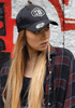 A black twill cap with a logo, worn by a model with long hair, standing in front of a graffiti background.