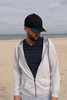A man wearing a grey zip-up hoodie and a black cap with a blue peak stands on a beach.