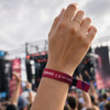 A hand wearing a vibrant pink and purple wristband, with a blurred concert crowd in the background and a stage visible.