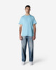 A man wearing a light blue Gildan Light Cotton T-shirt paired with faded jeans and white shoes, standing against a plain background.