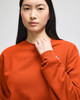 An adult crewneck sweatshirt in orange worn by a model, featuring a logo on the sleeve.