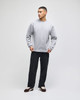 A grey crewneck sweatshirt with long sleeves, paired with dark trousers and casual shoes. The model stands against a plain background.