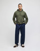 A man stands wearing an olive green pullover hooded sweatshirt and navy blue pants, with a logo visible on the hoodie.