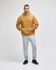 A hooded sweatshirt in a warm mustard colour paired with light grey sweatpants. The model is standing confidently.