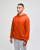 An orange pullover hooded sweatshirt with a front pocket and a logo on the sleeve. Styled with grey bottoms.