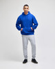 A blue pullover hooded sweatshirt with a front pocket, paired with grey sweatpants. The model stands confidently.