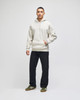 A light grey pullover hooded sweatshirt worn by a model, paired with dark trousers and shoes. The sweatshirt has a logo.