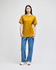 A model wearing a mustard yellow Champion adult short-sleeve T-shirt paired with blue jeans. The shirt features a logo.