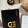 A black A5 weekly diary for 2026, featuring a logo and placed on a light surface with office accessories.